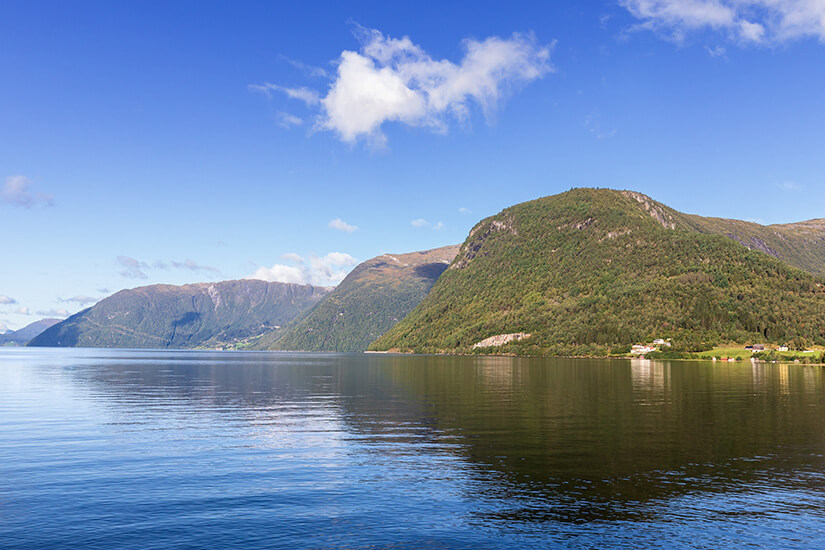 Zu erkennen ist ein ruhiger Fjord, umgeben von steilen, dicht bewaldeten Bergen. Das klare Wasser spiegelt die Landschaft und den blauen Himmel mit vereinzelten Wolken wider. Norwegen See