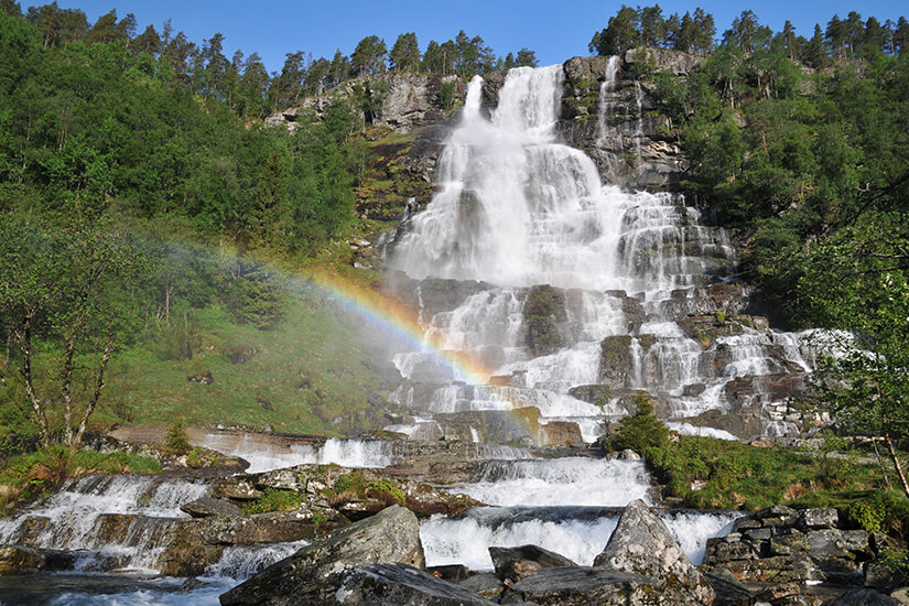 Zu erkennen ist ein mächtiger, mehrstufiger Wasserfall, der über felsige Kaskaden durch eine grüne Berglandschaft stürzt. Im aufgewirbelten Wassernebel hat sich ein deutlich sichtbarer Regenbogen gebildet. Nordfjordeid Wasserfall