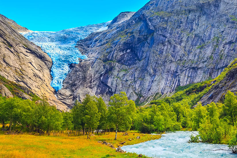 Zu erkennen ist eine eindrucksvolle Gletscherzunge zwischen steilen, felsigen Berghängen. Im Vordergrund fließt ein türkisfarbener Gletscherfluss durch eine grüne, von Bäumen bewachsene Landschaft. Norwegen Gletscher