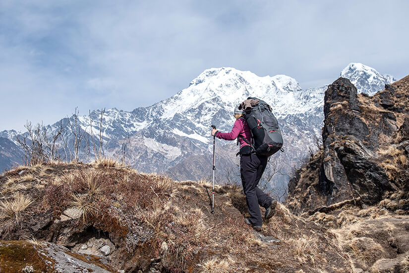 Mit großem Rucksack, Trekkingstock und Mütze steigt eine Wanderin in violetter Jacke über einen felsigen Grat mit trockenem Büschelgras. Vor ihr erhebt sich ein mächtiges, schneebedecktes Bergmassiv unter grau verhangenem Himmel, rechts ragen dunkle Felsnadeln auf. Packliste Peru Gebirge