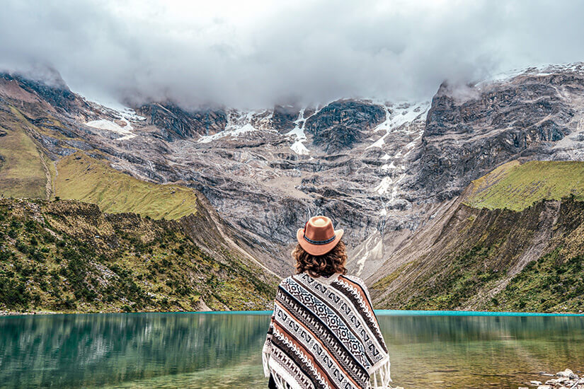 Von hinten gesehen sitzt ein Urlauber mit braunem Hut und gemustertem Poncho vor einem türkisgrün schimmernden Gletschersee und blickt auf die Bergkulisse. Packliste Peru Bergsee