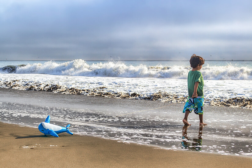 Barfuß steht ein kleiner Junge in grünem Shirt und bunter Badehose im auslaufenden Wasser und blickt auf die schäumende Brandung hinaus. Packliste Peru Strand