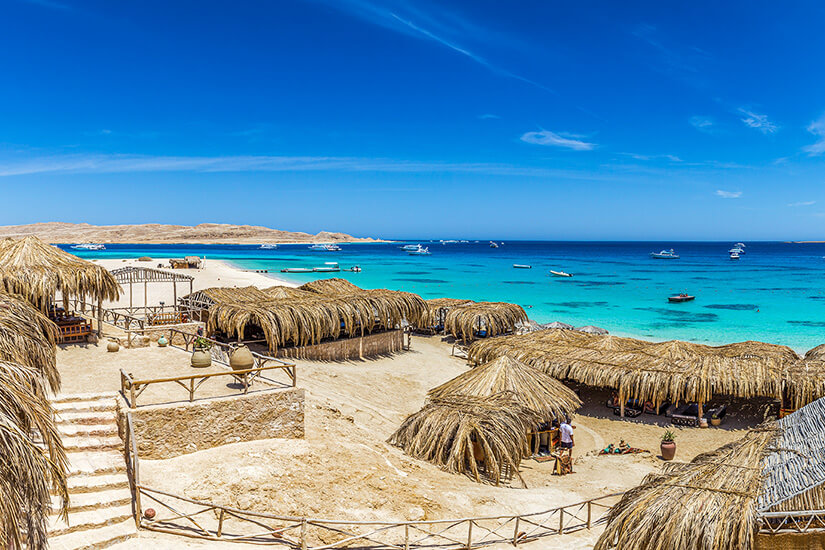 Strohgedeckte Sonnenschirme und gemütliche Sitzgelegenheiten auf Mahmya Beach vor Hurghada mit Blick auf das leuchtend türkisfarbene Meer. Im Hintergrund ankern Ausflugsboote in der geschützten Lagune der Giftun-Insel. Ägypten Strände Mahmya Beach