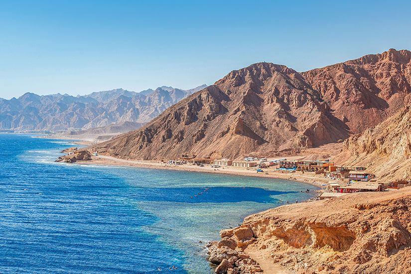 Blick über die schroffe Sinai-Küste bei Dahab, wo ockerfarbene Berge auf das tiefblaue Rote Meer treffen. Am Ufer reihen sich die typischen Beduinen-Cafés und kleinen Tauchbasen entlang des Küstenstreifens. Die Gegend rund um das Blue Hole gehört zu den legendärsten Tauchrevieren weltweit. Ägypten Strände Dahab