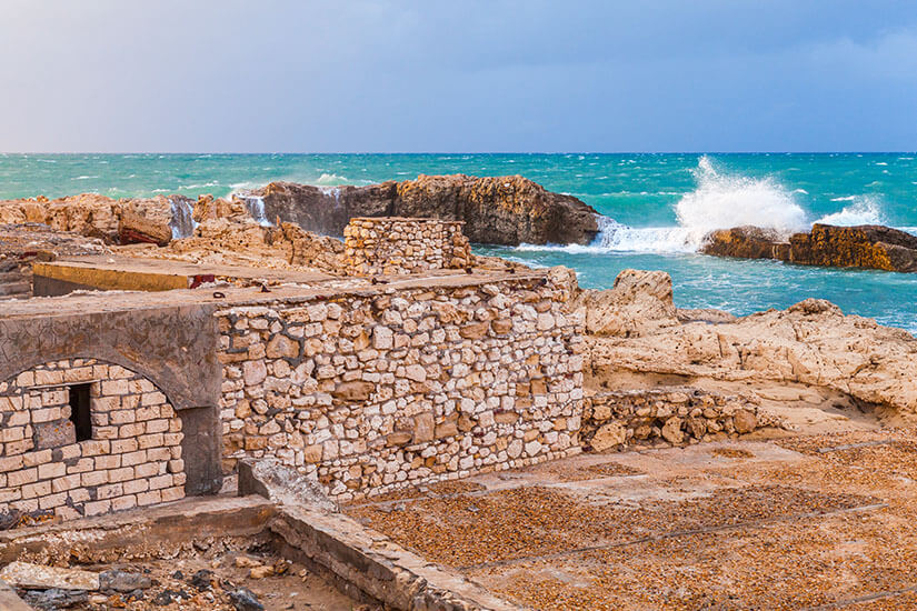 Historische Steinmauern am Ufer von Montazah Beach in Alexandria, wo antike Ruinen auf das türkisfarbene Mittelmeer treffen. Die Brandung bricht sich an den zerklüfteten Felsen und erzeugt eine dramatische Szenerie. Ägypten Strände Montazah Beach