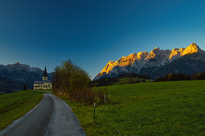 Eine schmale Asphaltstraße führt zwischen grünen Wiesen zu einer kleinen Kirche mit spitzem Turm auf einer Anhöhe. Gegenüber leuchtet eine schroffe Felskette im goldenen Abendrot, während der Himmel darüber bereits in tiefes Blau übergeht. Bischofshofen Buchbergkirche