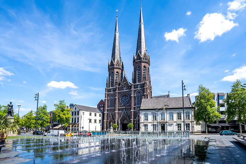 Zu erkennen ist eine neugotische Kirche mit zwei hohen, spitzen Türmen an einem weitläufigen Stadtplatz in Tilburg. Im Vordergrund befinden sich moderne Wasserspiele, während Bäume und historische Gebäude den Platz säumen. Tilburg Stadt