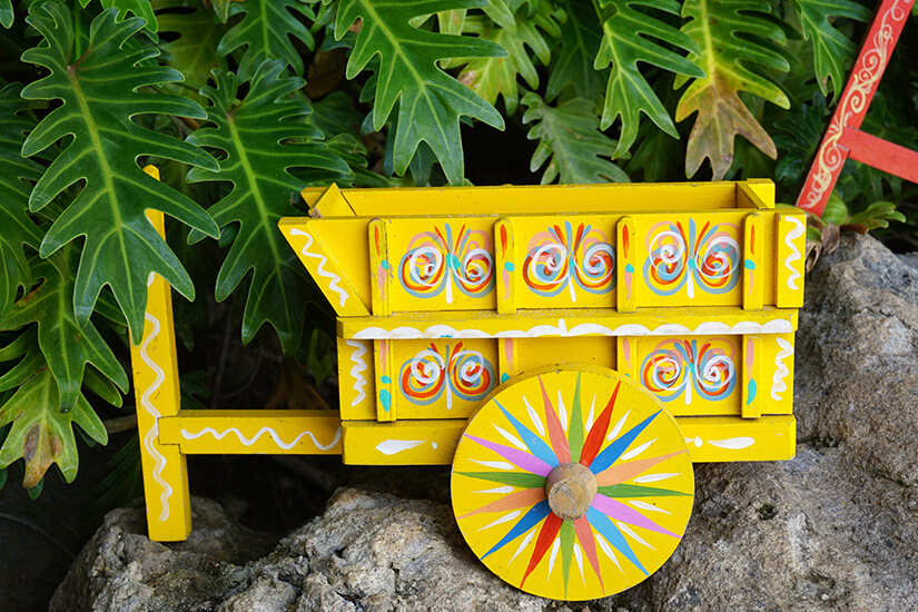 Zu erkennen ist ein kleines, leuchtend gelbes Modell eines traditionellen Ochsenkarrens mit farbenfrohen Ornamenten. Der kunstvoll bemalte Wagen steht vor üppigen grünen Blättern. Costa Rica Souvenirs Carreta