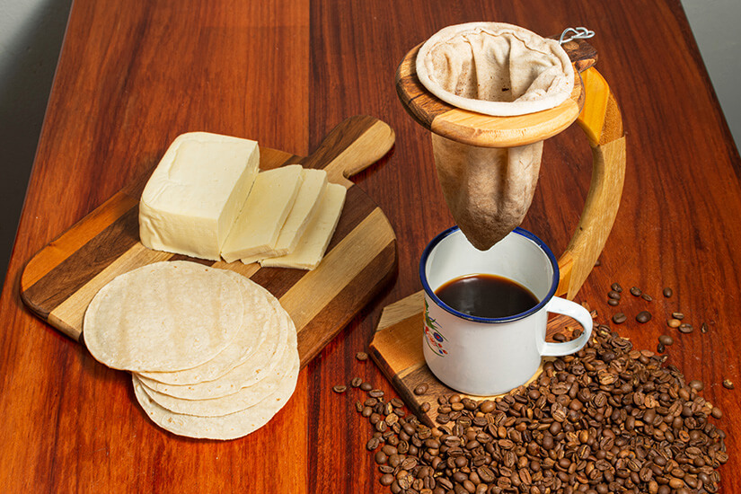Zu erkennen ist eine traditionelle Kaffeezubereitung mit einem Stofffilter über einer Tasse, umgeben von gerösteten Kaffeebohnen. Daneben liegen Tortillas und ein Stück heller Käse auf einem Holzbrett. Souvenirs Costa Rica Kaffeemaschine