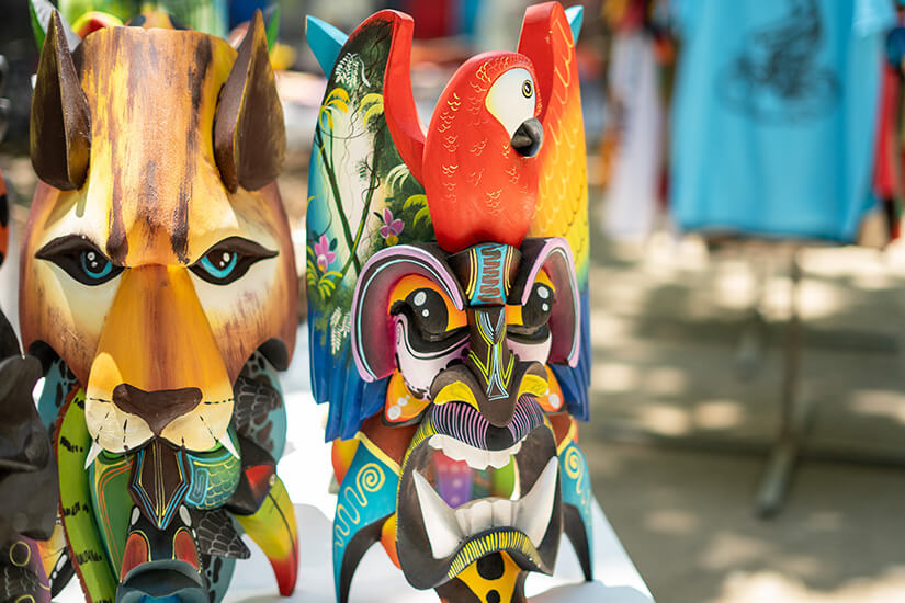 Zu erkennen sind farbenfrohe, kunstvoll bemalte Holzmasken mit Tiermotiven. Besonders auffällig sind ein Papagei sowie eine große Raubkatzenmaske, ergänzt durch weitere fantasievoll gestaltete Tierfiguren. Costa Rica Mitbringsel Masken