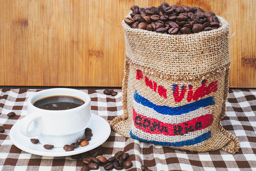 Zu erkennen ist eine Tasse schwarzer Kaffee neben einem Jutesack, der mit gerösteten Kaffeebohnen gefüllt ist. Der Sack trägt die Farben und den Schriftzug „Costa Rica“. Costa Rica Souvenirs Kaffee