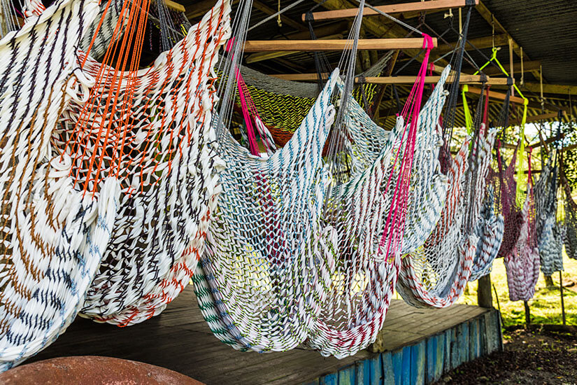 Zu erkennen sind zahlreiche handgeknüpfte Hängematten in unterschiedlichen Farben und Mustern. Sie hängen dicht nebeneinander an Holzbalken und vermitteln einen traditionellen, handgefertigten Eindruck. Costa Rica Geschenk Haengematte