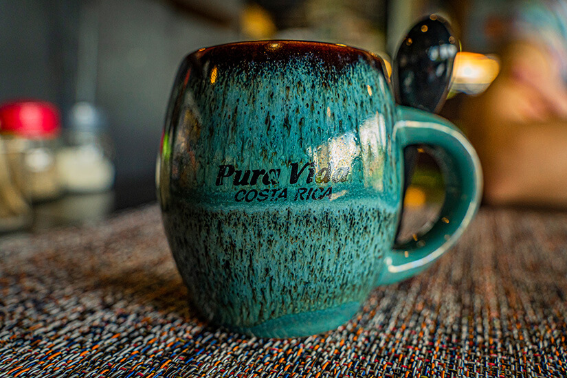 Zu erkennen ist eine türkisfarbene Keramiktasse mit der Aufschrift „Pura Vida Costa Rica“. In der Tasse steckt ein Löffel, während der Hintergrund unscharf gehalten ist. Costa Rica Andenken Pura Vida