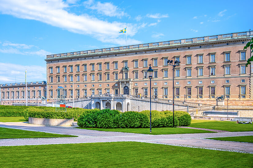 Das imposante, barocke Stockholmer Schloss mit seiner langgestreckten, beigefarbenen Fassade unter blauem Himmel. Auf dem Dach weht die schwedische Flagge. Im Vordergrund erstrecken sich gepflegte grüne Rasenflächen, akkurat geschnittene Buschreihen und gepflasterte Wege. Sehenswuerdigkeiten Schloss Stockholm Sehenswuerdigkeiten Schloss