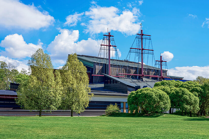 Das Vasa-Museum in Stockholm mit seinen charakteristischen, rot gestrichenen Schiffsmasten, die aus dem modernen Gebäudedach herausragen. Das Museum ist von gepflegten grünen Rasenflächen und dichten Laubbäumen umgeben unter einem blauen Himmel mit flockigen Sommerwolken. Stockholm Sehenswuerdigkeiten Vasa