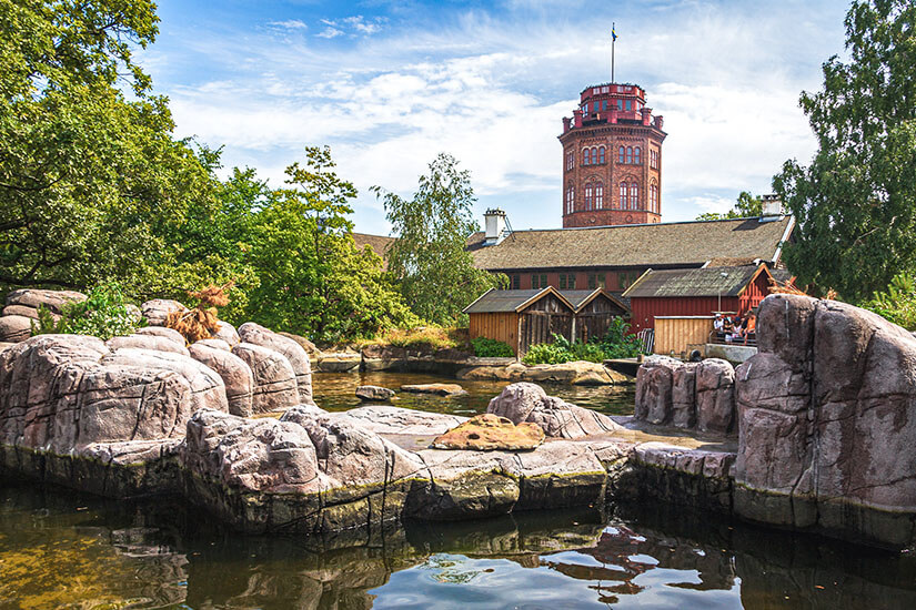 Ein Teich mit felsiger Kulisse im Freilichtmuseum Skansen in Stockholm. Im Hintergrund stehen traditionelle schwedische Holzhäuser und der markante rote Brendström-Aussichtsturm aus Backstein zwischen üppig grünen Bäumen unter leicht bewölktem Himmel. Stockholm Sehenswuerdigkeiten Skansen