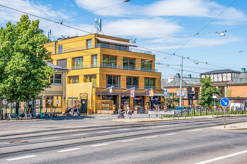 Das moderne, senfgelbe Gebäude des ABBA The Museum an einer Straße mit Straßenbahnschienen in Stockholm. Vor dem Gebäude stehen Passanten und E-Scooter, während grüne Bäume und weitere historische Häuser die Szenerie bei blauem Sommerhimmel umrahmen. Stockholm Sehenswuerdigkeiten ABBA