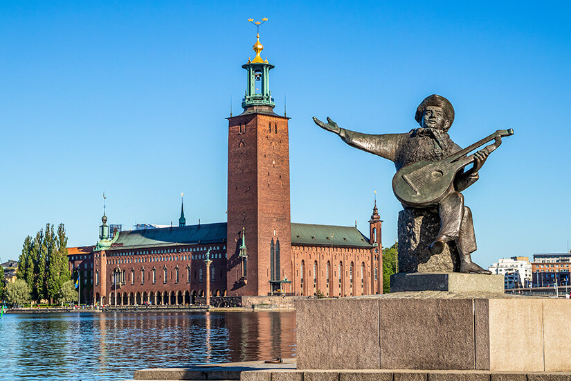 Eine Bronze-Skulptur des Dichters Evert Taube mit einer Laute im Vordergrund am Ufer des Mälaren-Sees. Im Hintergrund erhebt sich das imposante Stockholmer Rathaus (Stadshuset) aus rotem Backstein mit seinem markanten Turm und der goldenen Spitze vor strahlend blauem Himmel. Stockholm Sehenswuerdigkeiten Rathaus