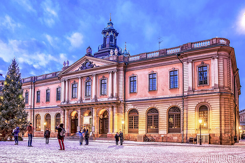 Das historische Börsengebäude am Stortorget-Platz in Stockholm, das das Nobelpreis-Museum beherbergt, in der Abenddämmerung. Die altrosa Fassade wird warm beleuchtet, während links ein großer, geschmückter Weihnachtsbaum auf dem leicht schneebedeckten Kopfsteinpflaster steht. Stockholm Sehenswuerdigkeiten Nobel Museum
