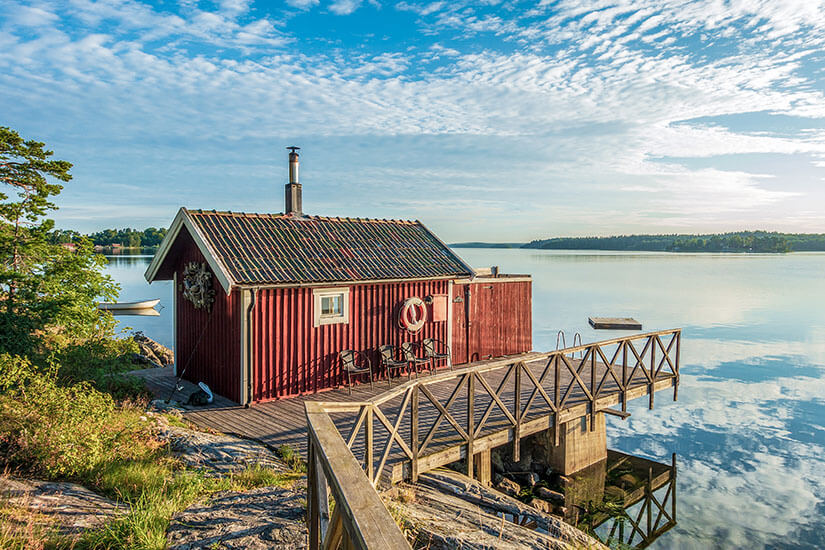 Ein klassisches, schwedenrotes Holzhaus mit Holzsteg auf Felsen direkt am ruhigen Wasser der Stockholmer Schären. Das spiegelglatte Wasser reflektiert den blau-weißen Wolkenhimmel. Stockholm Sehenswuerdigkeiten Schaeren