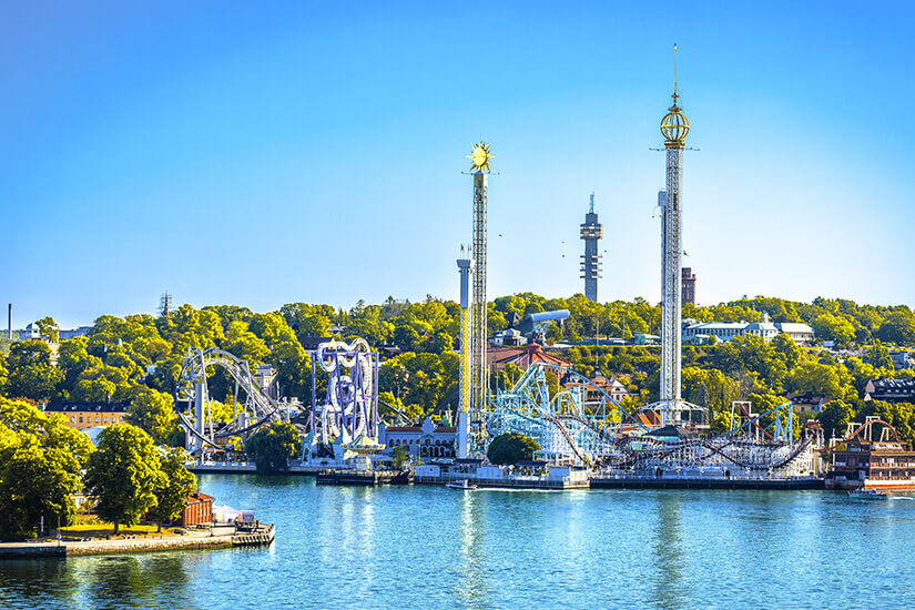 Blick über das blaue Wasser auf den Vergnügungspark Gröna Lund auf der Insel Djurgården in Stockholm. Zu sehen sind verschiedene Achterbahnen, ein hoher Kettenkarussell-Turm sowie ein Freefall-Tower vor einer Kulisse aus dichten, grünen Bäumen unter klarblauem Himmel. Stockholm Sehenswuerdigkeiten Groena Lund