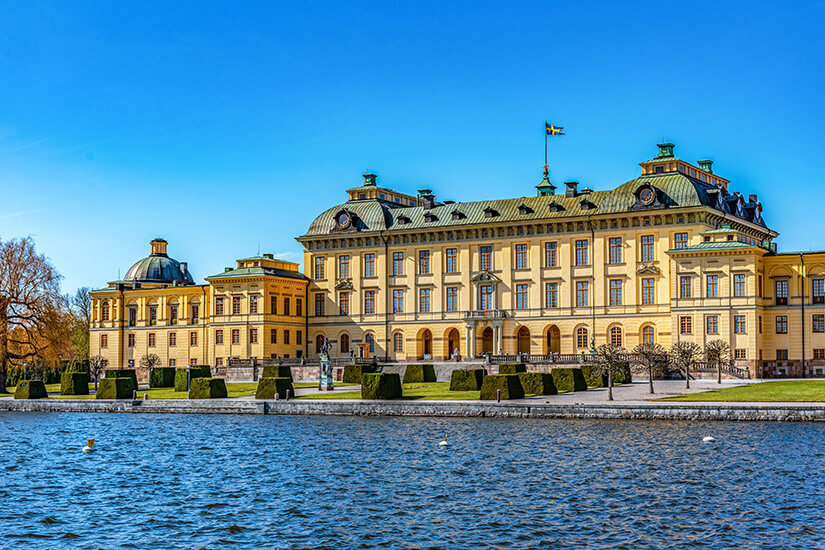 Das prachtvolle, gelb gestrichene Schloss Drottningholm mit seinen grünen Dächern am Ufer des Mälaren-Sees. Auf dem Dach weht die schwedische Flagge. Im Vordergrund glitzert das blaue Wasser, während im Schlossgarten akkurat geschnittene Hecken und Bäume stehen. Stockholm Sehenswuerdigkeiten Drottningholm
