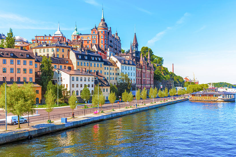 Panoramaansicht auf die bunten, historischen Gebäude von Södermalm entlang der Uferpromenade in Stockholm. Das tiefblaue Wasser glitzert in der Sonne, während spitze Türme und prachtvolle Backsteinfassaden den Hügel über der Uferstraße prägen unter einem klaren, blauen Himmel. Stockholm Sehenswuerdigkeiten Ausblick