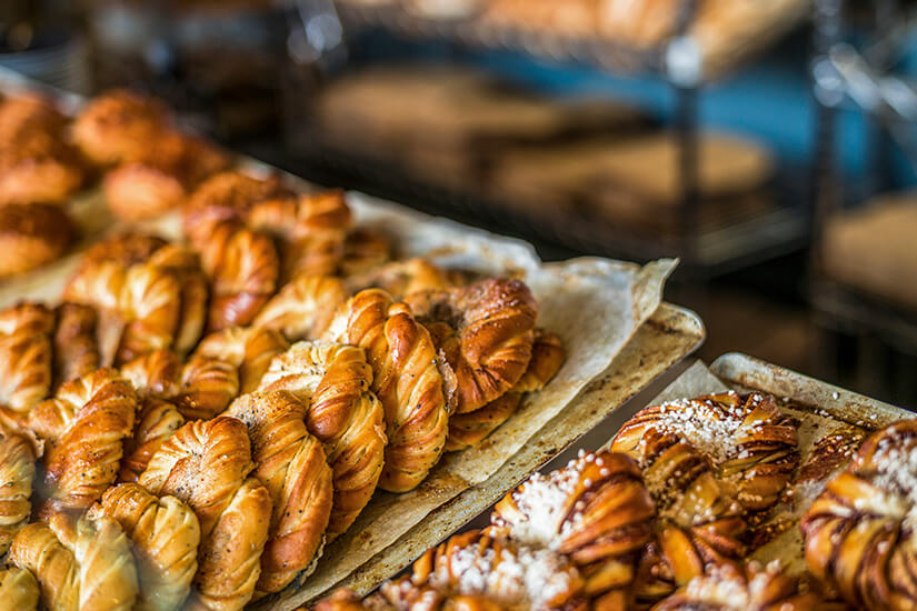 Frisch gebackene, golden gebräunte schwedische Zimtschnecken (Kanelbullar) und Kardamomschnecken liegen dicht an dicht auf Backblechen in einer Bäckerei. Einige der kunstvoll geknoteten Gebäckstücke sind mit Hagelzucker bestreut. Stockholm Sehenswuerdigkeiten Fika