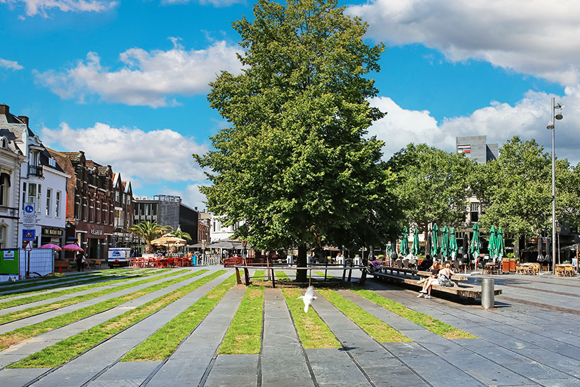 Zu erkennen ist ein weitläufiger Platz im Zentrum von Tilburg mit großen Bäumen, Grünstreifen und modernen Sitzgelegenheiten. Rundherum befinden sich Cafés, Restaurants und verschiedene Gebäude, während einige Menschen den sonnigen Platz nutzen. Tilburg Zentrum