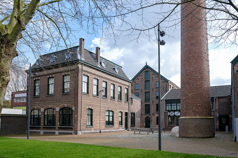 Zu erkennen ist ein historischer Backsteinkomplex mit großen Fenstern und einem markanten hohen Schornstein. Die ehemalige Industriearchitektur ist von einer gepflegten Grünfläche und gepflasterten Wegen umgeben. Tilburg Museum