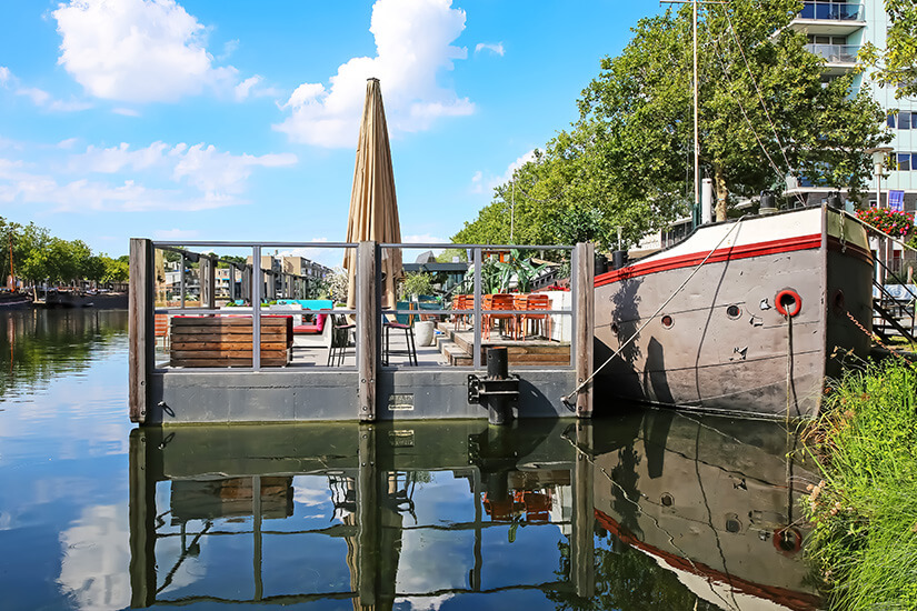 Zu erkennen ist ein großes, festgemachtes Schiff an einem ruhigen innerstädtischen Gewässer, daneben eine schwimmende Terrasse mit Tischen und Sitzgelegenheiten. Bäume und moderne Gebäude säumen das Ufer im Hintergrund. Tilburg Hafen