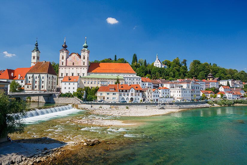 Die Stadt Steyr ist zu sehen. Eine Brücke führt über den Fluss. Das Wasser leuchtet grünblau, während am Ufer weiße Häuser mit roten Dächern und weitere Gebäude zwischen Bäumen aufragen. Steyr Oesterreich Willkommen