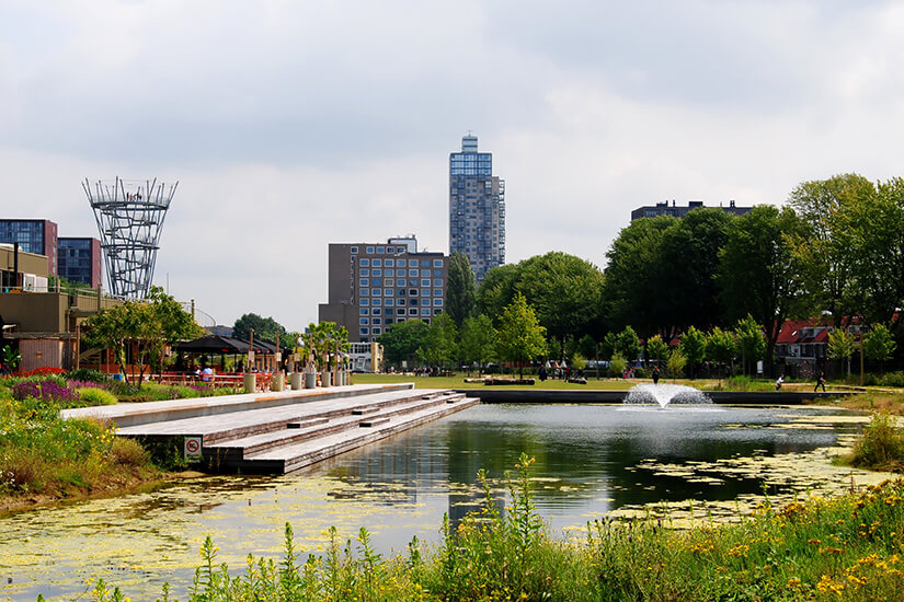 Zu erkennen ist ein moderner Stadtpark mit einem länglichen Teich, Springbrunnen und üppiger Begrünung. Im Hintergrund ragen mehrere Hochhäuser auf, während Wege und Sitzbereiche entlang des Wassers zum Verweilen einladen. Tilburg Park