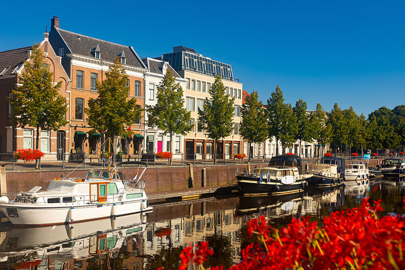 Zu erkennen ist eine malerische Uferpromenade mit historischen Gebäuden, Bäumen und mehreren festgemachten Booten. Das ruhige Wasser spiegelt die Häuser und Boote, während leuchtend rote Blumen im Vordergrund farbliche Akzente setzen. Niederlande Breda