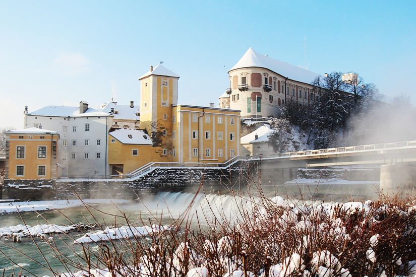 Die Dächer von Steyr und der Fluss sind im Winterkleide zu sehen. Am Ufer sind die Sträucher von Schnee bedeckt. Steyr Oesterreich Winter