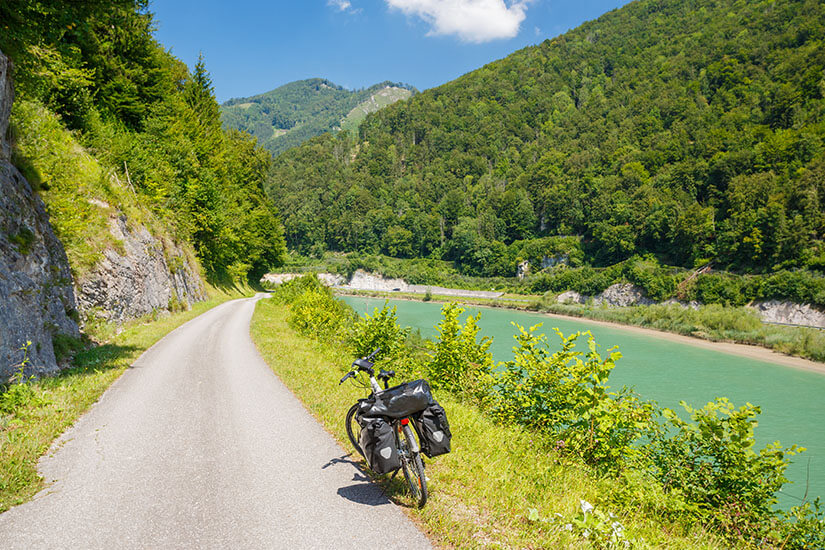Eine Radstrecke führt vorbei an grünen Hügeln und entlang eines türksifarbenen Flusses. Ein Fahrrad parkt am Ufer. Steyr Oesterreich Ennsradweg