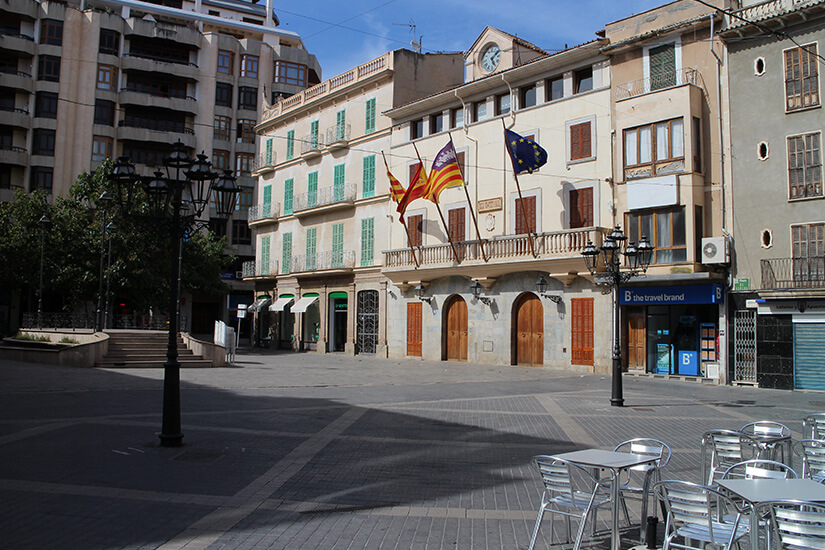 Der weitläufige, gepflasterte Hauptplatz Plaça d'Espanya in Inca auf Mallorca mit dem historischen Rathausgebäude im Zentrum. Am Rathaus hängen verschiedene Flaggen, darunter die spanische und die mallorquinische Flagge. Im Vordergrund stehen leere Cafétische und Stühle aus Metall im sonnigen Schattenbereich. Inca Mallorca Rathaus