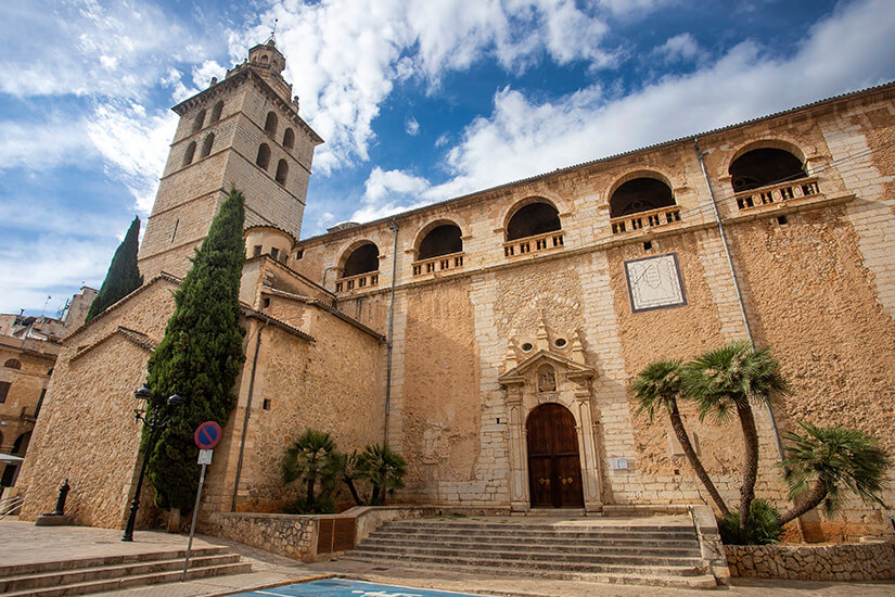 Die beeindruckende Barockkirche Es Església de Santa Maria Major in Inca auf Mallorca mit ihrer hellen Natursteinfassade und dem markanten Glockenturm. Eine breite Treppe führt zum hölzernen Eingangsportal, das von kleinen Palmen und Zypressen eingerahmt wird. Inca Mallorca Santa Maria Major