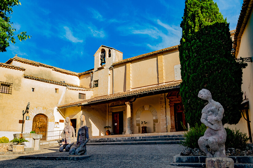 Der friedliche Innenhof des Klosters Monestir de Sant Bartomeu in Inca auf Mallorca mit hellen Sandsteinmauern und alten Rundbögen. Im Vorhof stehen abstrakte Steinskulpturen, historische Brunnen und hohe, dunkle Zypressen unter strahlend blauem Himmel. Inca Mallorca Sant Bartomeu