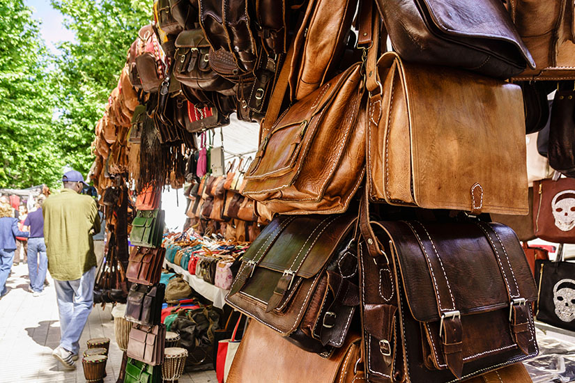 Nahaufnahme von handgefertigten braunen und dunklen Ledertaschen und Umhängetaschen an einem Marktstand im Freien. Im Hintergrund flanieren Besucher entlang der Marktstände im schattigen Freibereich. Inca Mallorca Ledertaschen