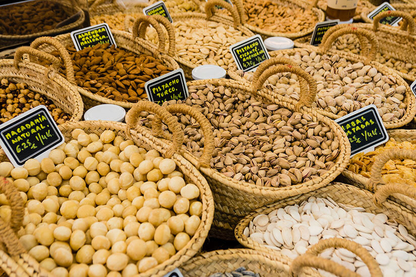 Verschiedene geflochtene Körbe voller Nüsse und Kerne, darunter Macadamianüsse, Mandeln und Pistazien, auf einem traditionellen Marktstand in Inca. Kleine Kreidetafeln beschriften die Sorten und Preise auf Spanisch. Inca Mallorca Markt