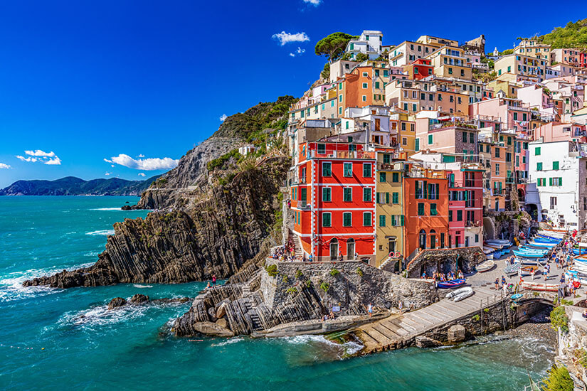 Zu erkennen ist Riomaggiore an der italienischen Ligurischen Küste mit dicht gestaffelten, farbenfrohen Häusern an einem steilen Felshang. Am kleinen Hafen liegen Boote, während das türkisfarbene Meer an die zerklüftete Felsküste brandet. Riomaggiore Stadt