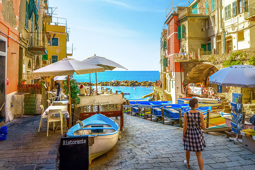 Zu erkennen ist eine schmale Gasse in Riomaggiore, die zwischen farbenfrohen Häusern direkt zum kleinen Hafen und zum Meer führt. Zahlreiche Boote, Sonnenschirme und ein Restaurant prägen die lebhafte mediterrane Atmosphäre. Riomaggiore Gassen