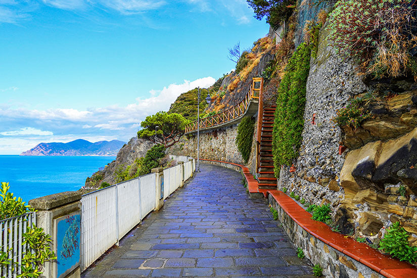 Zu erkennen ist ein gepflasterter Küstenweg, der direkt an einer steilen Felswand entlangführt. Links eröffnet sich ein weiter Blick auf das tiefblaue Meer und die bergige Küstenlandschaft. Italien Via Amore