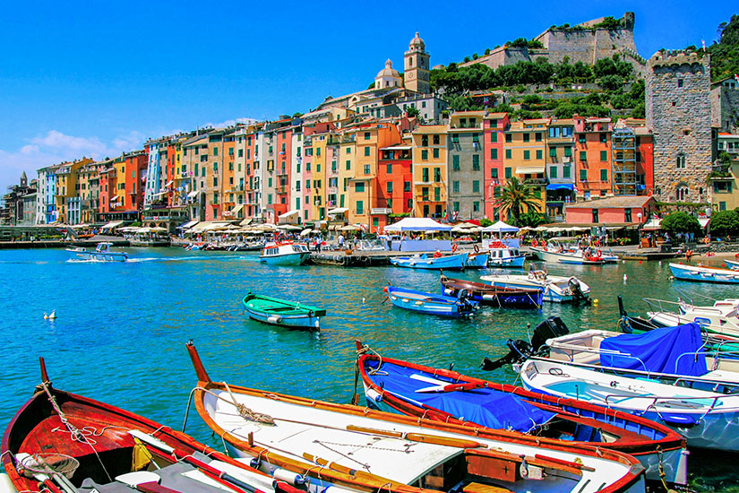 Zu erkennen ist der malerische Hafen von Portovenere mit zahlreichen Booten und einer Reihe farbenfroher Häuser direkt am Wasser. Oberhalb des Ortes erheben sich eine Kirche, historische Befestigungsanlagen und ein markanter Steinturm. Italien Porto Venere