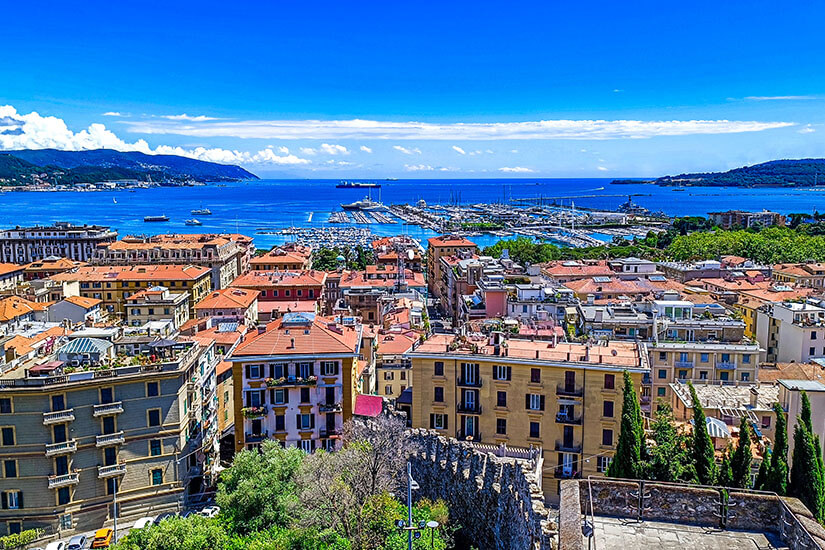 Zu erkennen ist ein weitläufiger Blick über La Spezia mit dicht bebauten Stadtvierteln und roten Ziegeldächern. Im Hintergrund erstrecken sich der große Yachthafen und die tiefblaue Bucht, eingerahmt von grünen Hügeln. Italien La Spezia