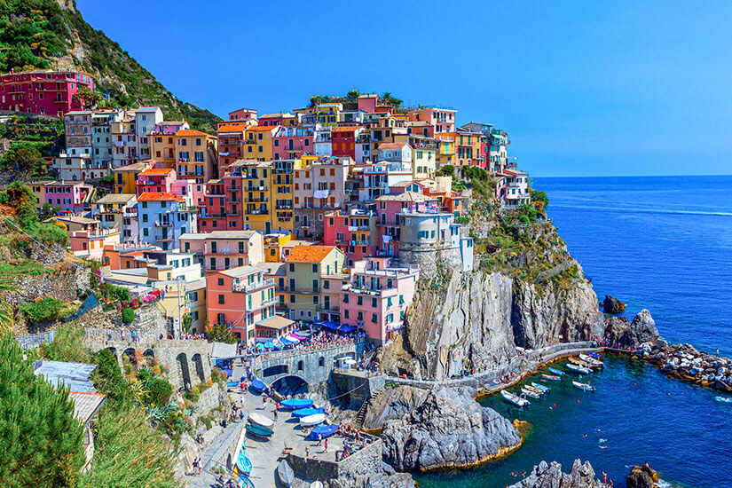 Zu erkennen ist Manarola mit seinen dicht an den steilen Felsen gebauten, farbenfrohen Häusern. Unterhalb des Ortes liegt eine kleine felsige Bucht mit Booten, während sich dahinter das tiefblaue Mittelmeer erstreckt. Italien Manarola