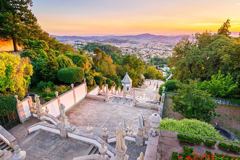 Von oben fällt der Blick auf die barocke Treppenanlage des Heiligtums Bom Jesus do Monte bei Braga, gesäumt von weißen Mauern, steinernen Statuen und üppigem Grün. Top Tagestrips Porto Braga