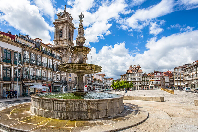 Im Vordergrund plätschert ein mehrstöckiger Steinbrunnen auf dem weitläufigen Largo do Toural im Herzen von Guimarães, dahinter ragt der Glockenturm der Kirche São Pedro in den Himmel. Top Tagestrips Porto Guimaraes