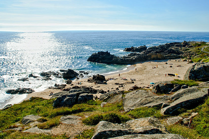 Zwischen dunklen Felsformationen schmiegt sich eine kleine, geschützte Sandbucht an der Atlantikküste bei Vila do Conde, an der vereinzelte Badegäste die Sonne genießen. Top Tagestrips Porto Vila do Conde